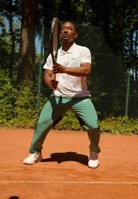 Tennisser in een witte polo met groen Nike-logo en groene trainingsbroek; houdt een tennisracket vast op een gravelbaan. Zonovergoten achtergrond.