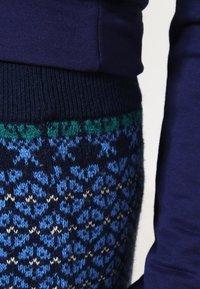 Primo piano di una persona che indossa un maglione blu scuro e una gonna di maglia blu con motivi, con dettagli verdi e bianchi in vita.
