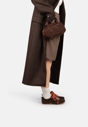 Borsa in suede marrone con dettagli in metallo dorato, tenuta in una mano, abbinata a un lungo cappotto marrone, pantaloni corti beige, calzini bianchi e sneakers scure.