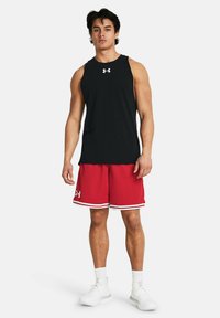 Top sin mangas negro y pantalones cortos deportivos rojos con ribetes blancos, que presentan el logo de Under Armour. El modelo lleva zapatillas blancas.