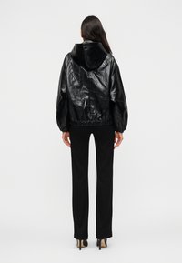 Γυναίκα που φοράει μαύρο γυαλιστερό μπουφάν με κουκούλα, μαύρο παντελόνι ίσια γραμμή και μαύρα μυτερά παπούτσια με τακούνια, στέκεται με την πλάτη στραμμένη προς τα εμπρός.