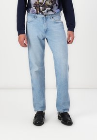 Jeans din denim de un albastru deschis, cu un design drept, cinci buzunare și un finisaj uzat, purtați cu o camasă cu imprimeu grafic cu mânecă lungă și pantofi negri.