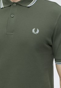 Πράσινο υφασμάτινο κοντομάνικο πόλο με λευκό κεντημένο λογότυπο στεφάνης δάφνης και διπλή λευκή ρίγα στον γιακά και στις άκρες των μανικιών.