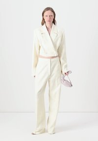 Blazer blanco recortado con doble botonadura y pantalones a juego de corte ajustado. Tejido suave con una sutil textura. La modelo sostiene un pequeño bolso de lavanda.