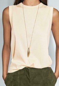 Donna che indossa un top beige senza maniche e pantaloni verde oliva, con una lunga collana di pendente d'oro che pende sopra la maglietta.