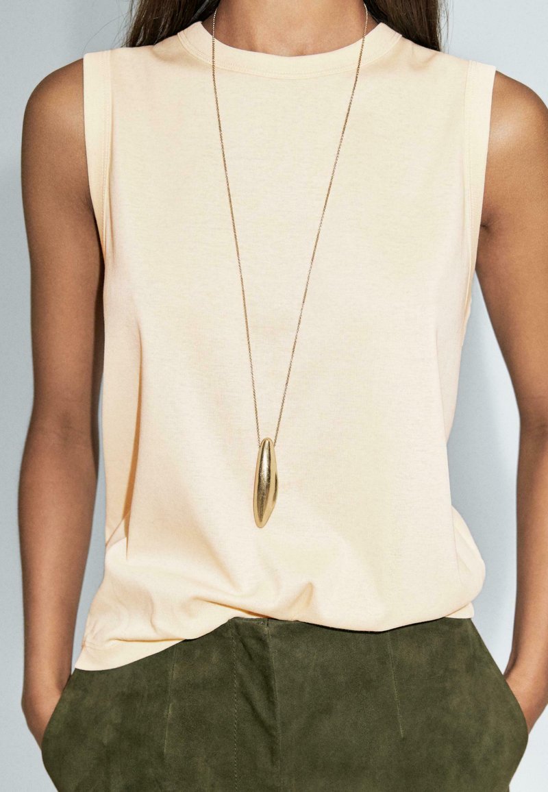 Donna che indossa un top beige senza maniche e pantaloni verde oliva, con una lunga collana di pendente d'oro che pende sopra la maglietta.