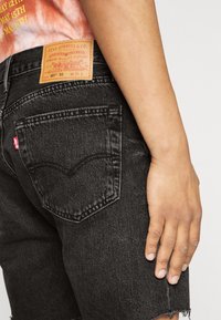 Shorts de mezclilla negra con dobladillo desgastado, con un parche trasero de cuero marrón claro grabado con los detalles de la marca y una etiqueta roja en el bolsillo trasero.
