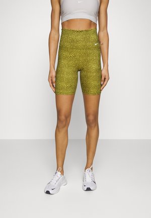 Kobieta ubrana w zielone, teksturowane, rowerowe szorty z wysokim stanem, szary sportowy biustonosz i białe sportowe buty, stojąca na jednolitym tle.