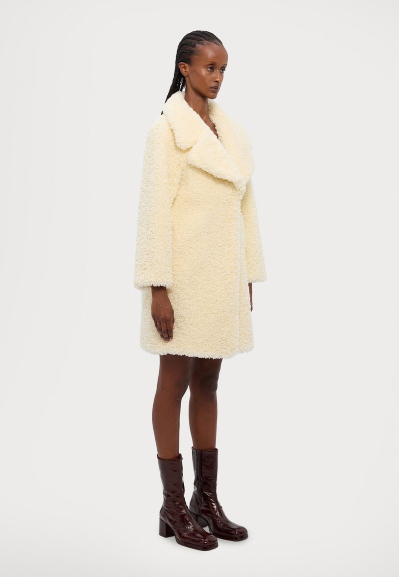 Cappotto in finta pelliccia di un morbido color crema, con un ampio colletto e una silhouette ampia, dotato di maniche lunghe e lunghezza fino a metà coscia. Stivali lucidi marroni al ginocchio.