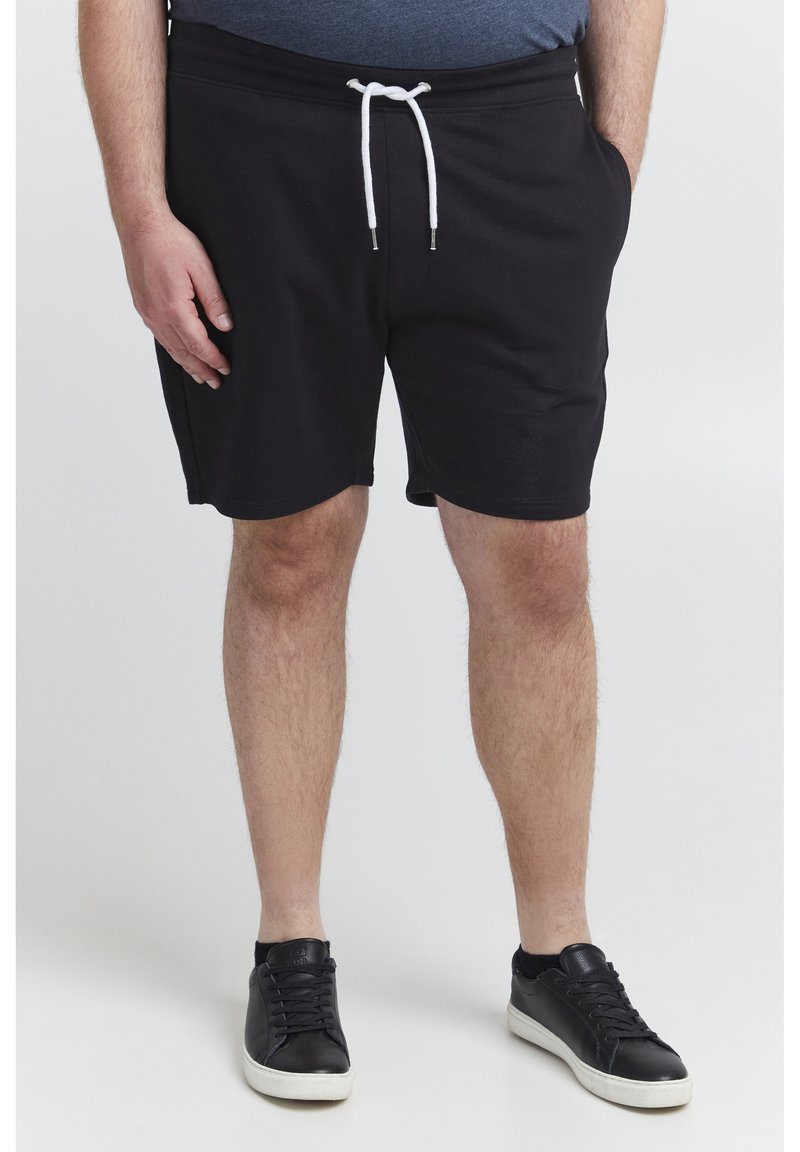 Mann iført svarte trening-shorts med snøring, mørkeblå skjorte og svarte sneakers med hvite såler, står med en hånd i lommen.