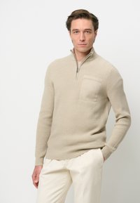 Beiger Strickpullover mit einem Viertelreißverschluss-Kragen, gerippter Textur und einer Brusttasche. Kombiniert mit hellen Hosen.