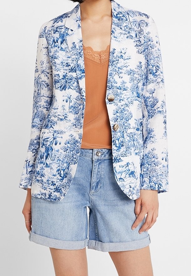 Blazer floreale blu e bianco con un design sartoriale, chiusura a un bottone e maniche arrotolate, abbinato a un top beige e shorts di denim.