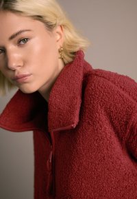 Pulover roșu din fleece cu guler înalt și închidere cu fermoar pe jumătate. Prezintă un material texturat și o croială lejeră. Accente aurii din metal.