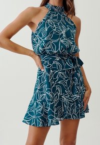 Turkos halterneck-klänning med vitt lövmönster, elastisk midja och volang fåll. Tillverkad av lättviktig tyg och har en knytbar midjedetalj.