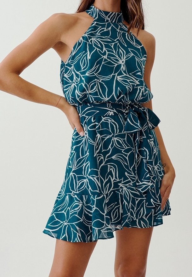 Turkos halterneck-klänning med vitt lövmönster, elastisk midja och volang fåll. Tillverkad av lättviktig tyg och har en knytbar midjedetalj.