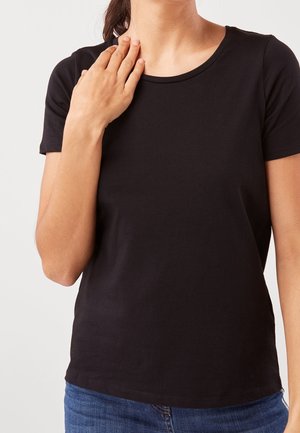 Femme portant un t-shirt noir à manches courtes et un jean bleu, la main droite reposant sur son épaule gauche devant un fond uni.