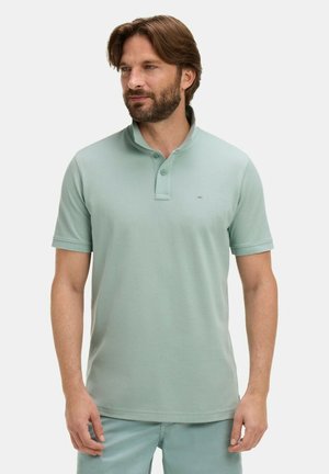 Homme avec une barbe portant un polo à manches courtes vert clair et un pantalon assorti, debout devant un fond blanc uni.