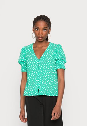 Blusa verde de manga curta com bolinhas brancas, decote em V e fecho com botões. Tecido texturizado; mangas franzidas. Calças pretas visíveis.