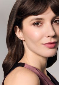 Brunetka w małych złotych kolczykach w kształcie kółek, z delikatnymi pomadkami, wypolerowaną cerą i sukience z ciemnego i jasnego materiału.