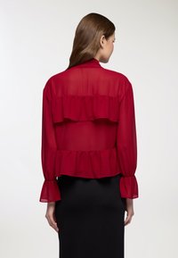 Blusa rossa trasparente con volant a strati, maniche lunghe con polsini a campana e una silhouette fluida. Indossata sopra una gonna nera.