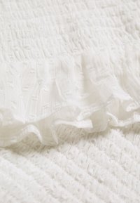 Bílá látka s nařasenou strukturou a volánkem. Materiál se jeví jako lehký a má jemné embosované detaily.