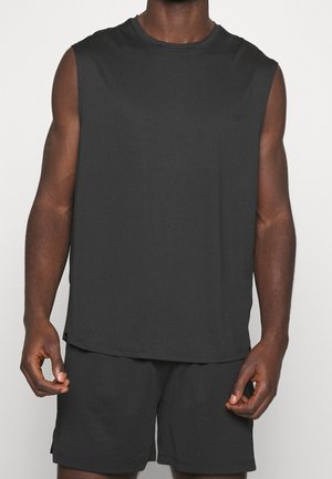 Mann trägt ärmelloses schwarzes Sportshirt und schwarze Shorts, steht vor einem weißen Hintergrund, Arme entspannt an den Seiten.