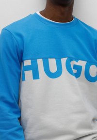 Helles blaues Sweatshirt mit einem dunkleren blauen oberen Bereich und fettem weißen "HUGO"-Text auf der Vorderseite. Aus weichem Baumwollmaterial hergestellt.