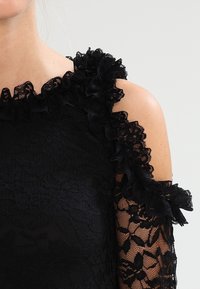 Svart spetsblus med volangdetaljer på axlarna, genomskinliga ärmar och ett texturerat blommönster i hela tyget.