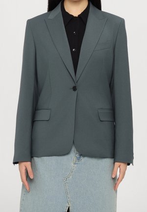 Kvinde iført en mørkegrøn blazer med enkel knap, sort skjorte med krave og en lyseblå denim nederdel med slidse foran.