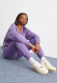 Fialová mikina a odpovídající tepláky vyrobené z měkké tkaniny. Krémové tenisky s růžovými akcenty. Model sedící na šedém koberci.