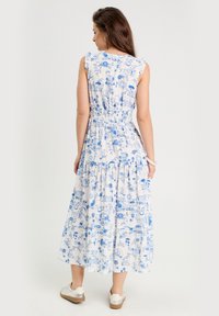 Robe maxi blanche avec motif floral bleu, manches courtes volantées, et taille élastique, avec jupe à volants et ourlet à la cheville.