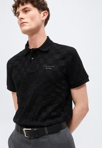 Νεαρός άνδρας που φοράει μαύρο καρό πόλο με το λογότυπο "Porsche BOSS", γκρι παντελόνι και μαύρη ζώνη, στέκεται μπροστά σε απλό φόντο.