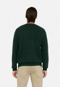 Dunkelgr�üner Strickpullover mit rundem Ausschnitt und gerippten Bündchen. Enthält Farbspritzer in verschiedenen Farben für Textur. Wird über hellen Hosen getragen.