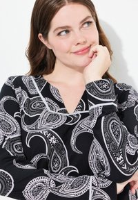 Musta paita, jossa on paisley-kuvio valkoisena ja harmaana, V-päänauhalla ja pitkillä hihoilla. Kangas näyttää sileältä ja siinä on hieman kiiltoa.