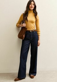 Top dolcevita giallo, jeans a gamba larga blu scuro a vita alta, cintura marrone e borsa a tracolla marrone, con scarpe marroni slip-on.