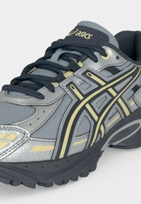 Γκρι και μαύρο αθλητικό παπούτσι Asics με κίτρινες λεπτομέρειες και μαύρα κορδόνια, που δείχνει λεπτομερείς πλευρικές και μπροστινές όψεις.