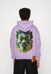 Felpa lavanda con un grande grafico di un volto verde e nero sul retro. Presenta un cappuccio con cordino e un'etichetta arancione sull'orlo.
