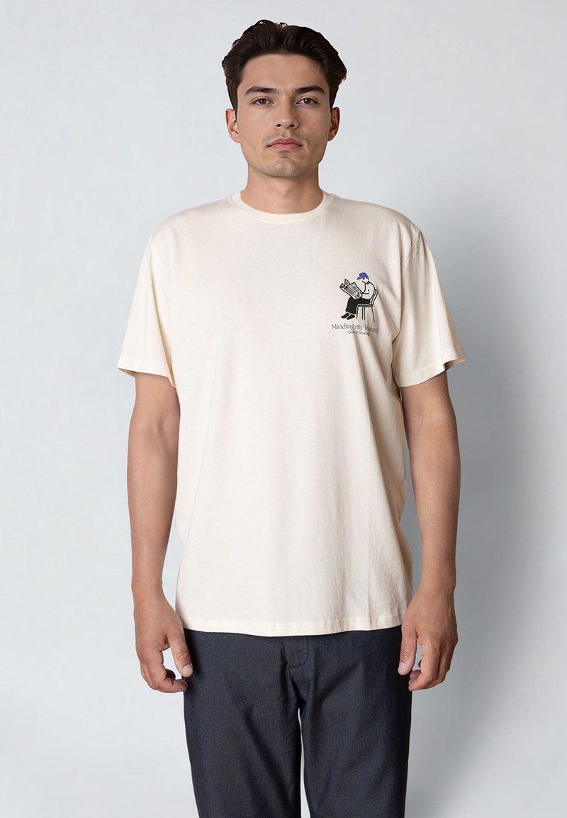 Κρεμ βαμβακερό T-shirt με κοντά μανίκια, διακοσμημένο με εικόνα χαρακτήρα που διαβάζει εφημερίδα, συνοδευόμενο από κείμενο στο στήθος.