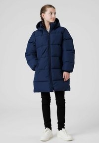 Marinblå puffjacka med huva, med vadderad design och dragkedja fram. Bärs över svarta leggings och vita sneakers.