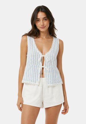 Donna con top senza maniche a righe azzurro chiaro e bianco con laccetti davanti e shorts bianchi a vita alta in piedi davanti a uno sfondo semplice.