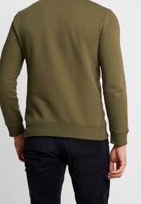 Sudadera verde oliva de manga larga, con puños y dobladillo acanalados. Textura suave, diseño ajustado. Combinada con pantalones oscuros de corte slim.