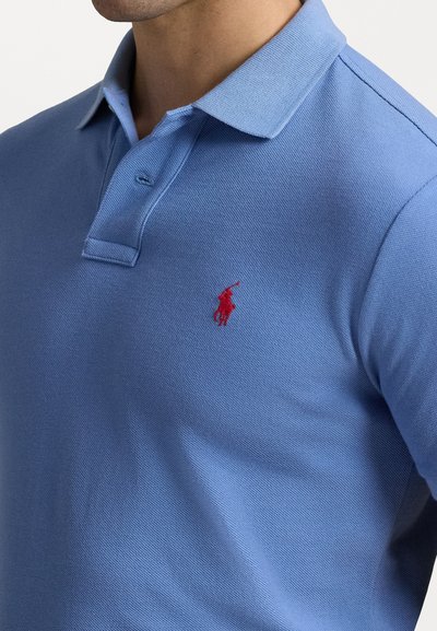 Polo de color azul claro hecho de una tela texturizada, con una tapeta de tres botones y un logo bordado en rojo en el lado izquierdo del pecho.