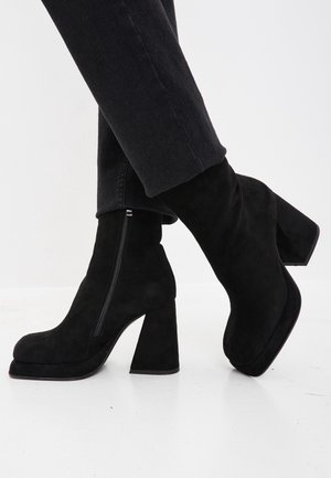 Bottines en daim noir à talons épais avec fermetures éclair latérales portées avec un pantalon noir sur fond blanc.