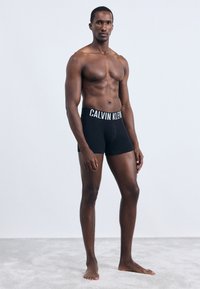 Fekete pamut boxer alsók fehér "CALVIN KLEIN" logós derékkal, testhez simuló fazonban, sima textúrával, amelyet egy mezítelen felsőtestű modell visel.