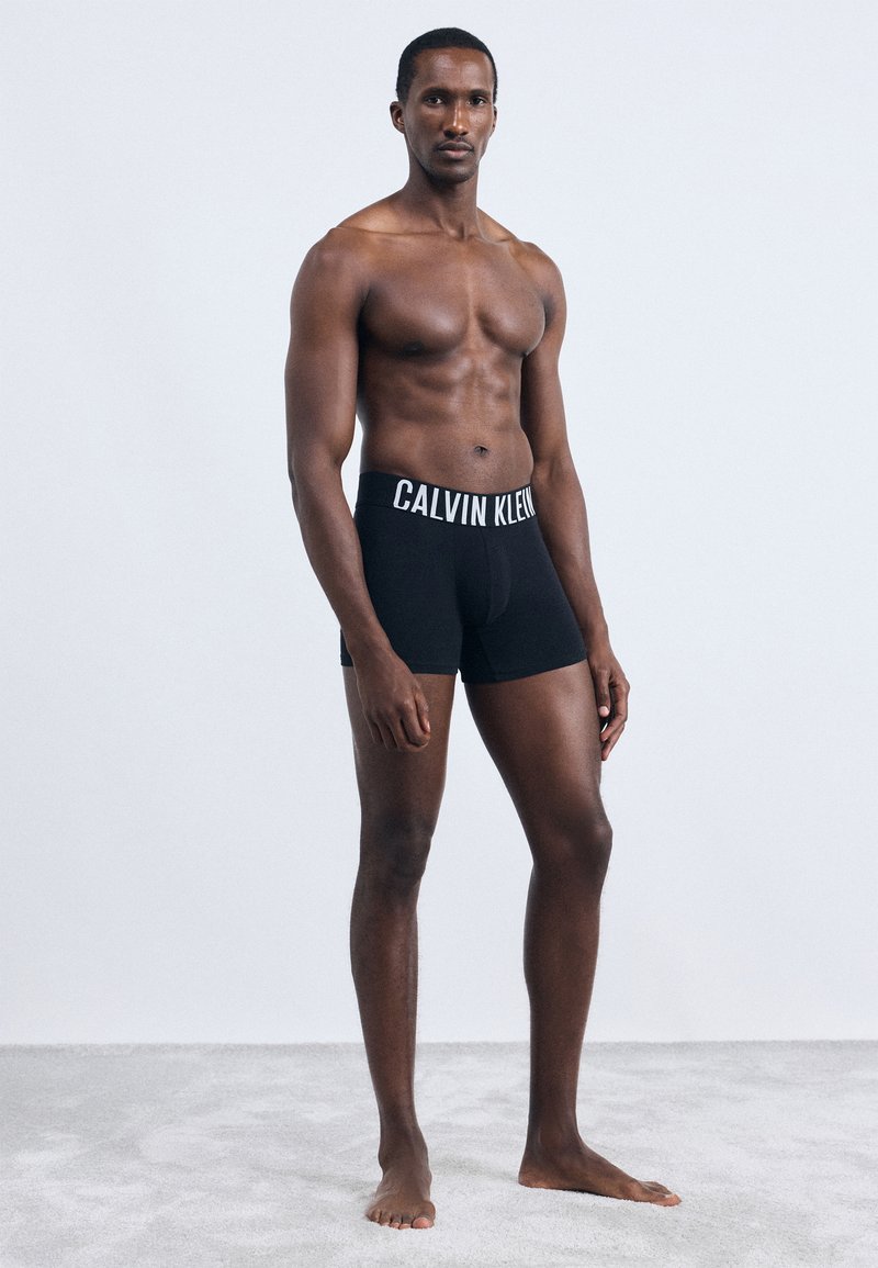 Fekete pamut boxer alsók fehér "CALVIN KLEIN" logós derékkal, testhez simuló fazonban, sima textúrával, amelyet egy mezítelen felsőtestű modell visel.