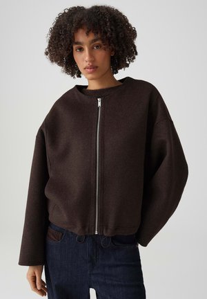 Femme aux cheveux bouclés portant une veste zippée marron foncé et un jean bleu foncé, debout devant un fond uni.