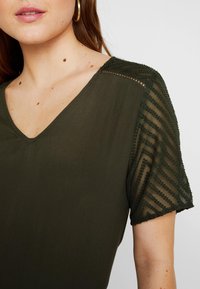 Olivengrøn top med V-hals og korte ærmer. Ærmet har struktureret, gennemsigtigt stof med et chevron mønster. Glat kropstof.