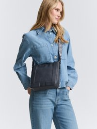 Czarna skórzana torebka na ramię o teksturowym wykończeniu, z wzorzystym paskiem i geometrycznymi akcentami, w połączeniu z dżinsową koszulą i dżinsami.