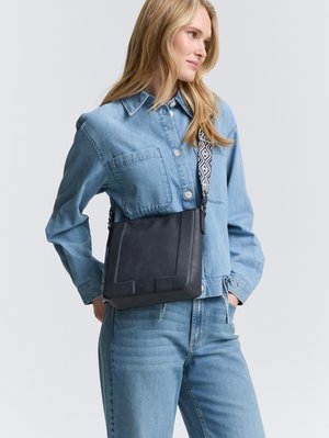 Schwarze Ledertasche zum Überwerfen mit strukturiertem Finish, versehen mit einem gemusterten Gurt und geometrischen Akzenten, kombiniert mit einem Jeanshemd und einer Jeans.