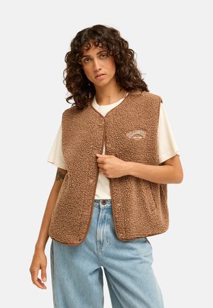 Femme aux cheveux bouclés portant une veste polaire marron sur un t-shirt blanc et un jean bleu clair, debout devant un fond uni.